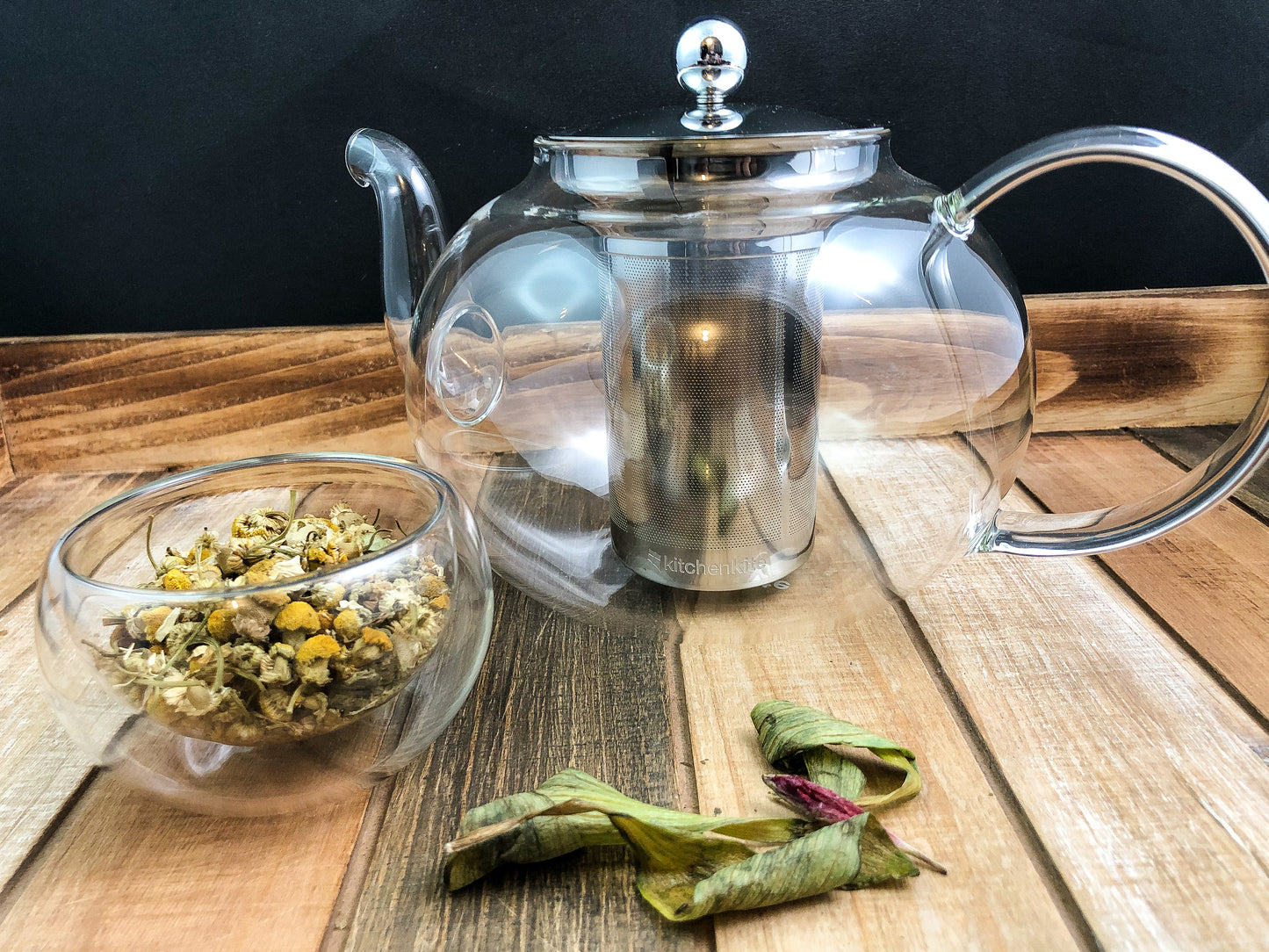clear glass tea pot with a clear small glass bowl filled with dried chamomile in left forefront and green leaves on a wooden table with a black background