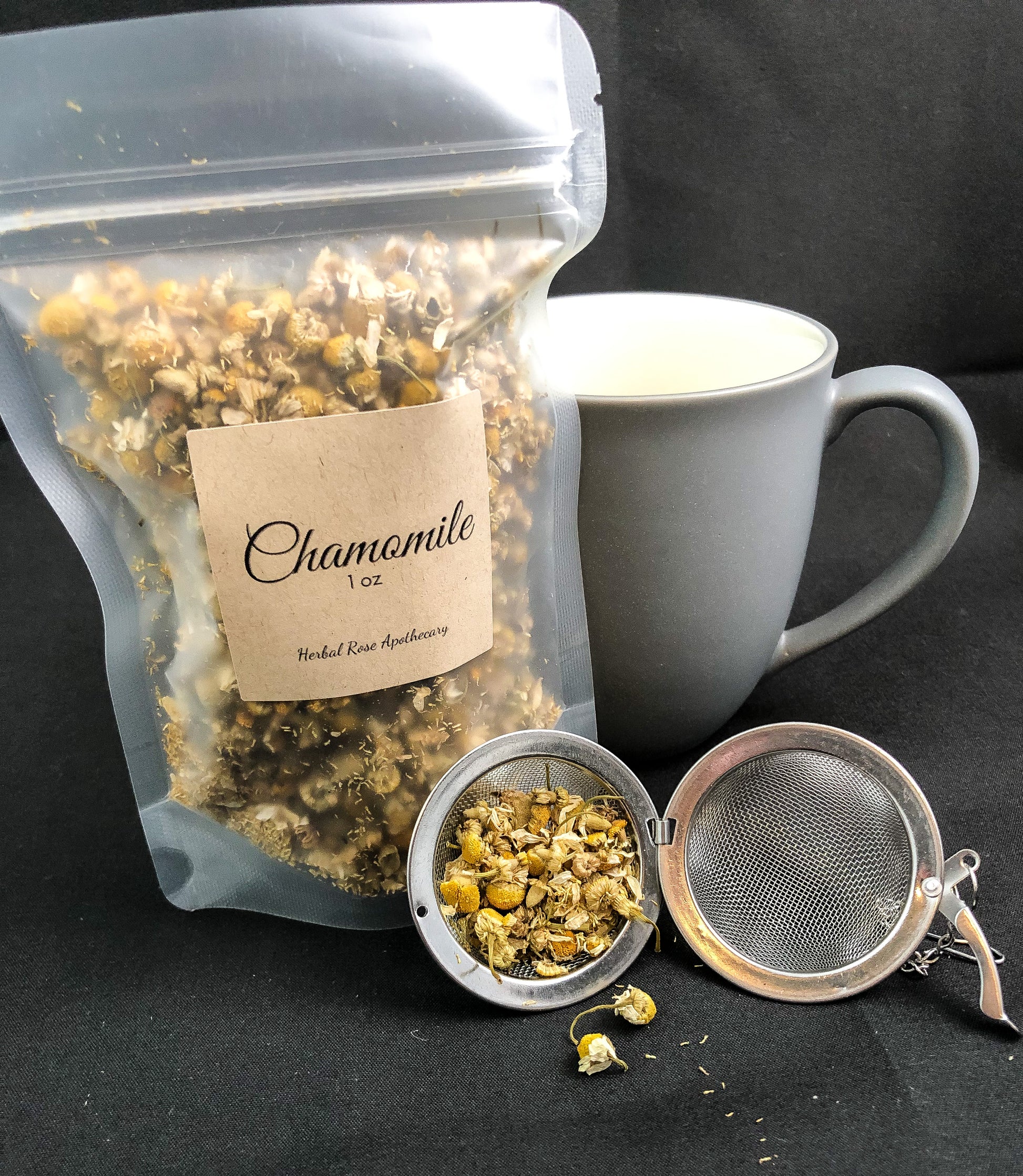 image of 1oz dried chamomile in clear plastic bag with a an open mesh tea strainer and chamomile inside and falling out, along with a grey mug next to bag and a black background