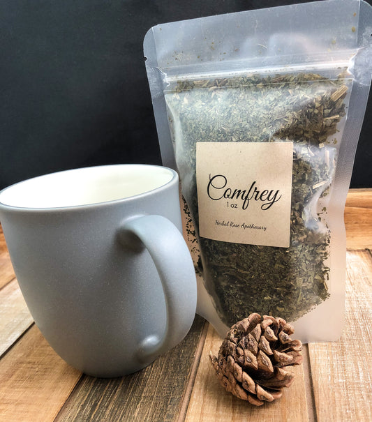 dried comfrey in 1oz clear bag next to a pine cone and grey mug with black background and items on a wooden table top