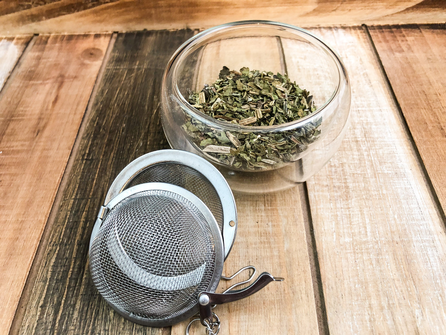 image of dried comfrey in a clear glass cup next to a mesh tea infuser with a wooden table as a background