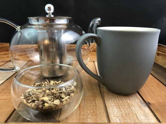 clear glass teapot with grey mug to the right and clear glass bowl of dried echinacea root in forefront with black background on a wooden table
