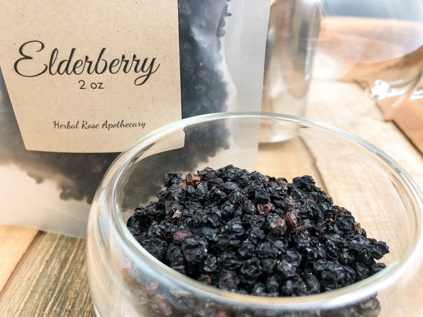 dried elderberry upclose in a clear glass cup next to a 2oz clear bag filled with elderberries on a wooden table