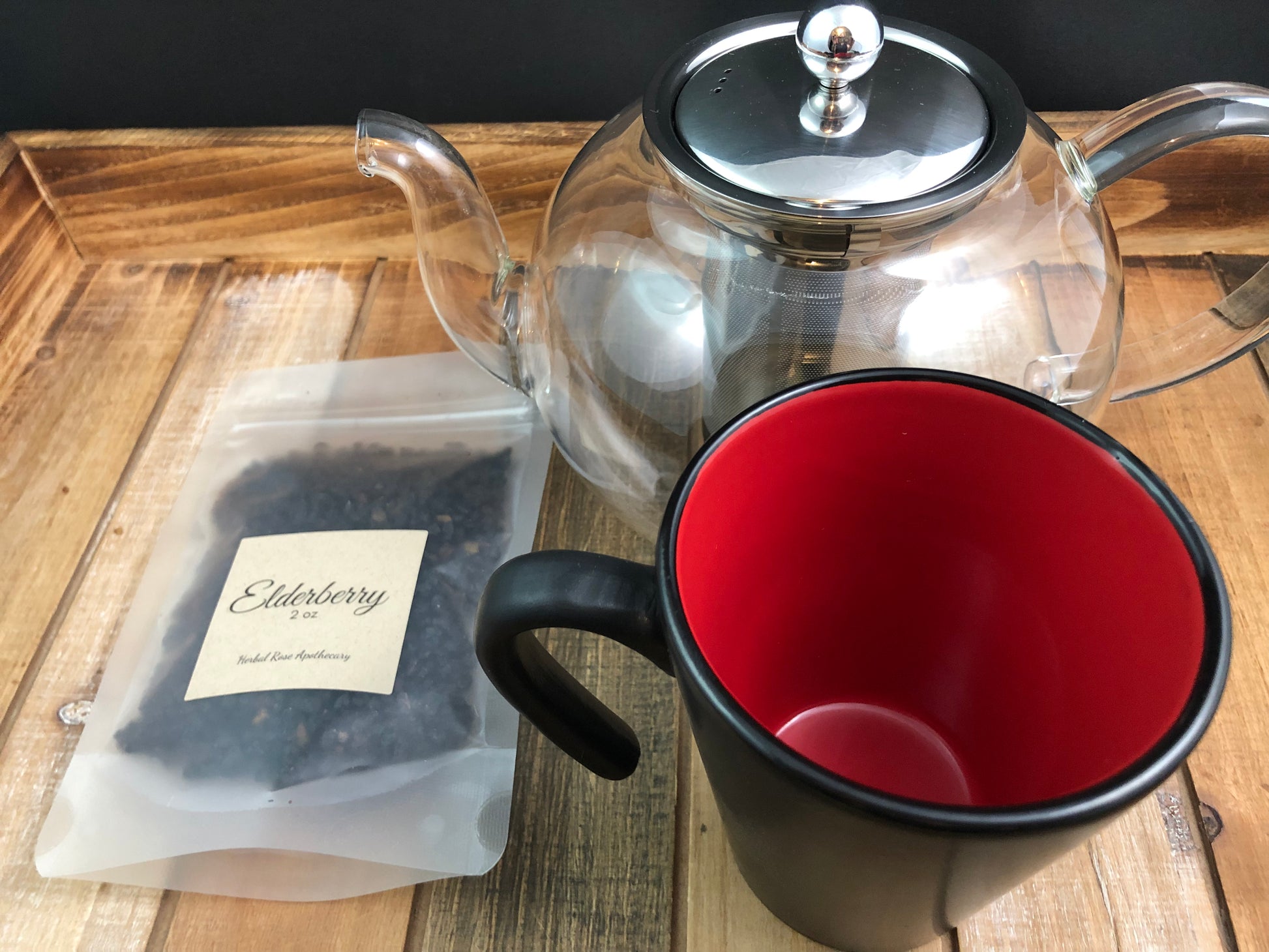 dried elderberry in a 2oz clear bag next to a clear tea pot and black and red mug with a black background and items on a wooden tray
