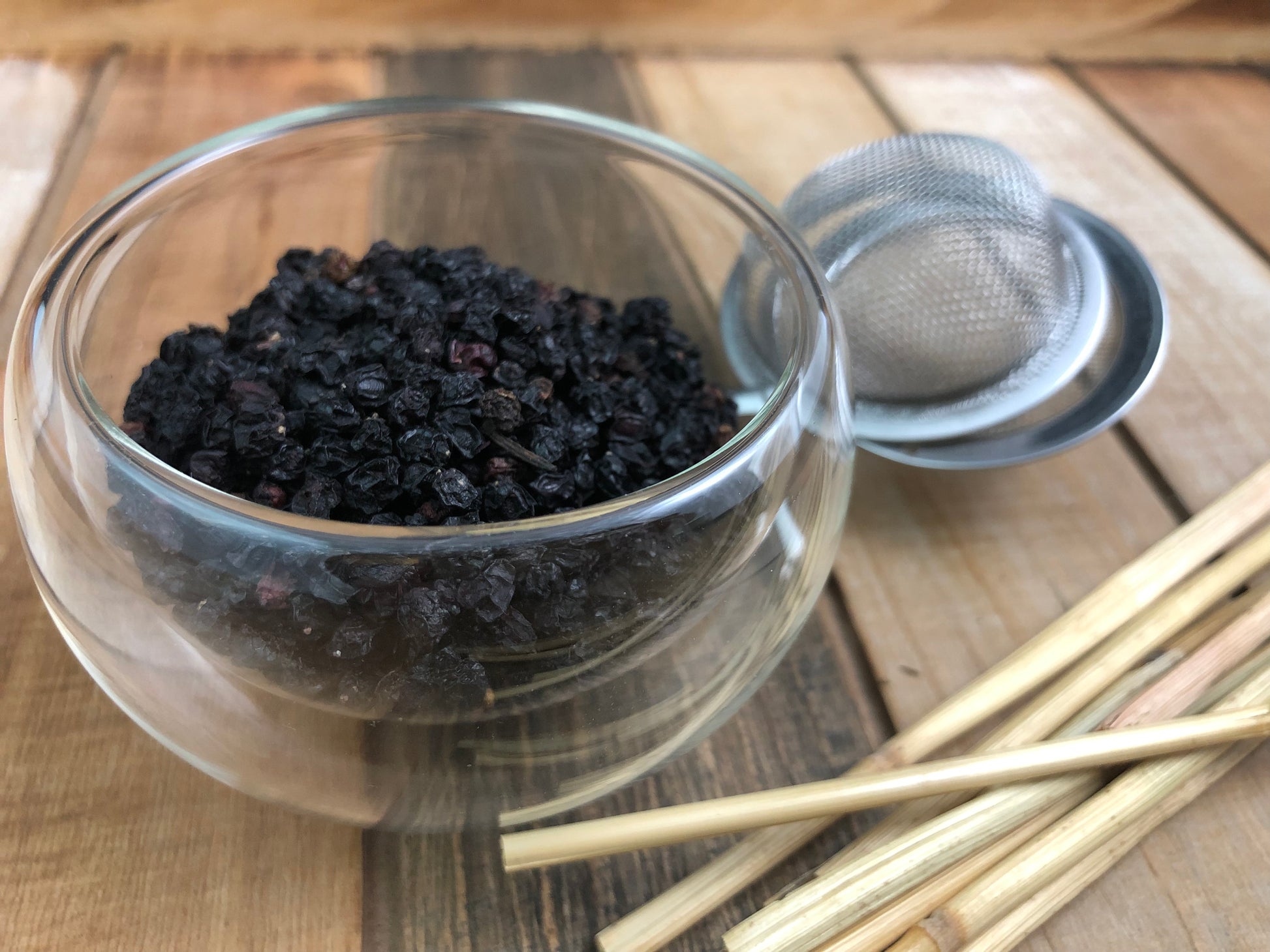 upclose image of dried elderberries in a clear glass cup next to a mesh tea infuser and wooden sticks on a wooden tray table