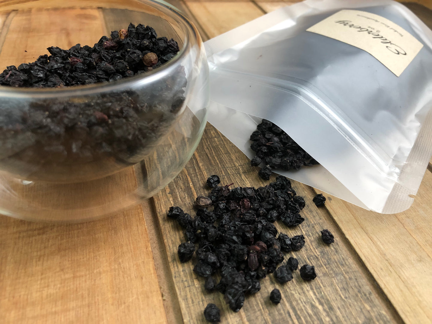 image of dried elderberries spilling out of clear bag next to a clear glass cup with dried elderberries, on a wooden table