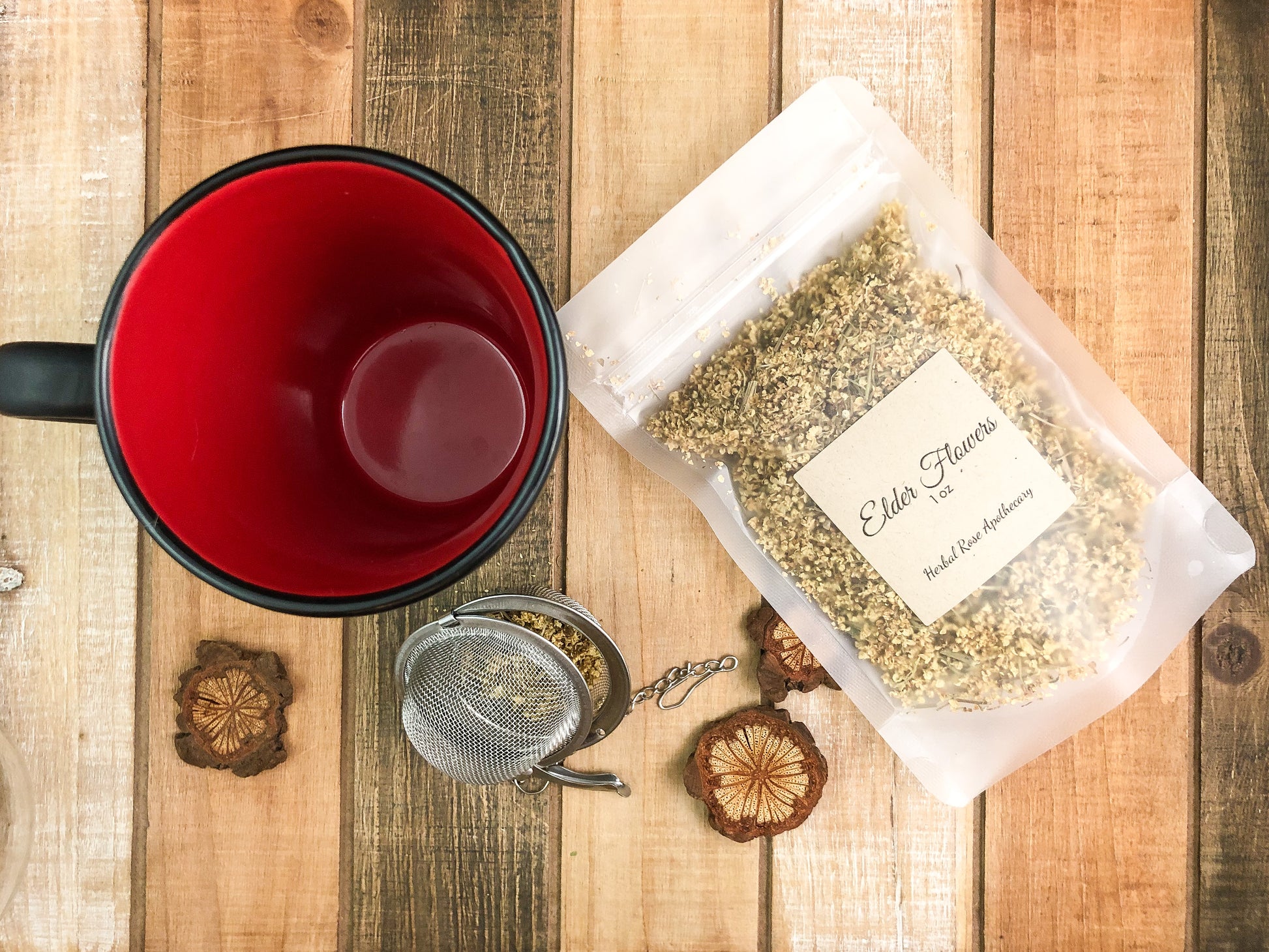 Ariel view of dried elderflowers in a clear bag laying flat next to a black mug  with red inside and mesh tea strainer, with wooden chips scattered around all on a wooden background
