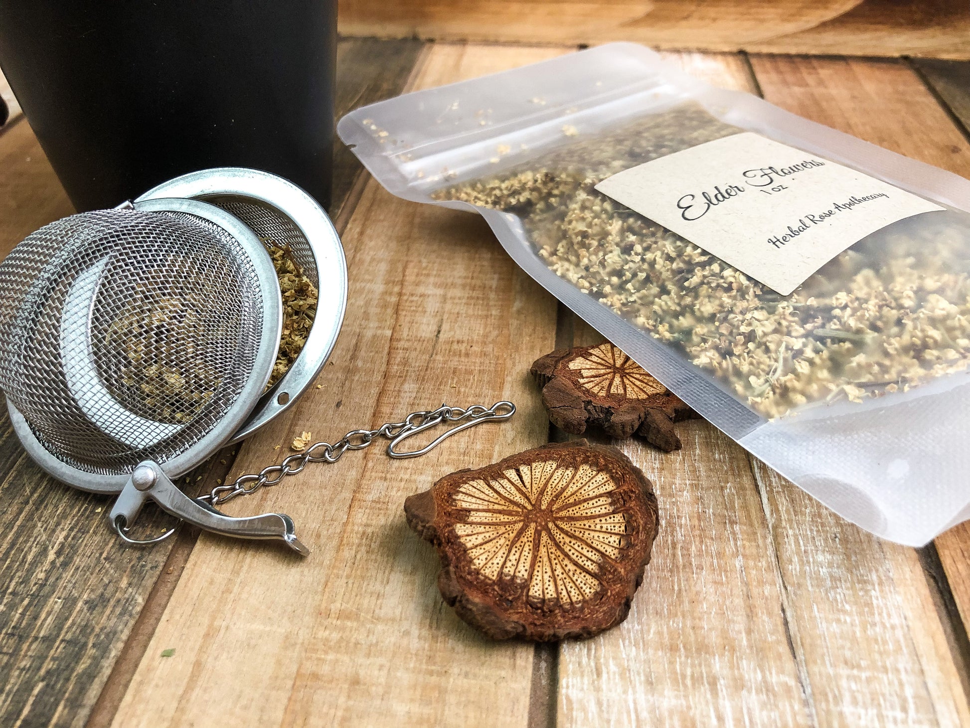 dried elderflowers in a clear bag laying flat next to a black mug and mesh tea strainer, with wooden chips scattered around all on a wooden background
