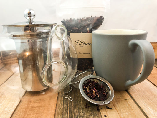 image of dried hibiscus in clear 2oz bag next to a grey coffee mug and a clear teapot  and mesh tea infuser, all items on a wooden table with white background