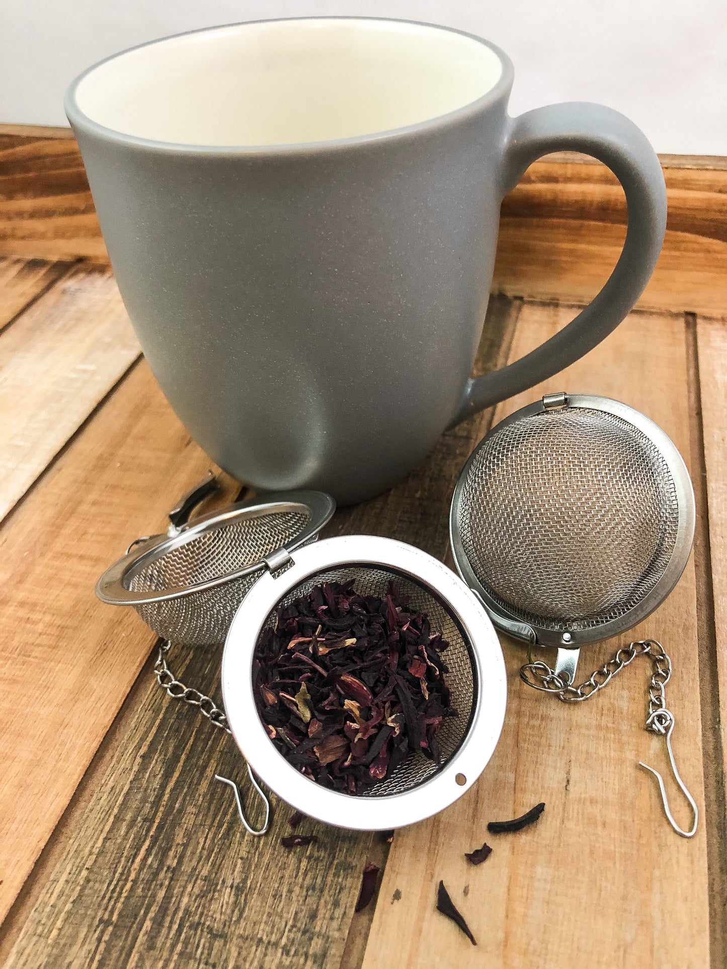 dried hibiscus in a mesh tea infuse next to a grey coffee mug on a wooden table with a white background