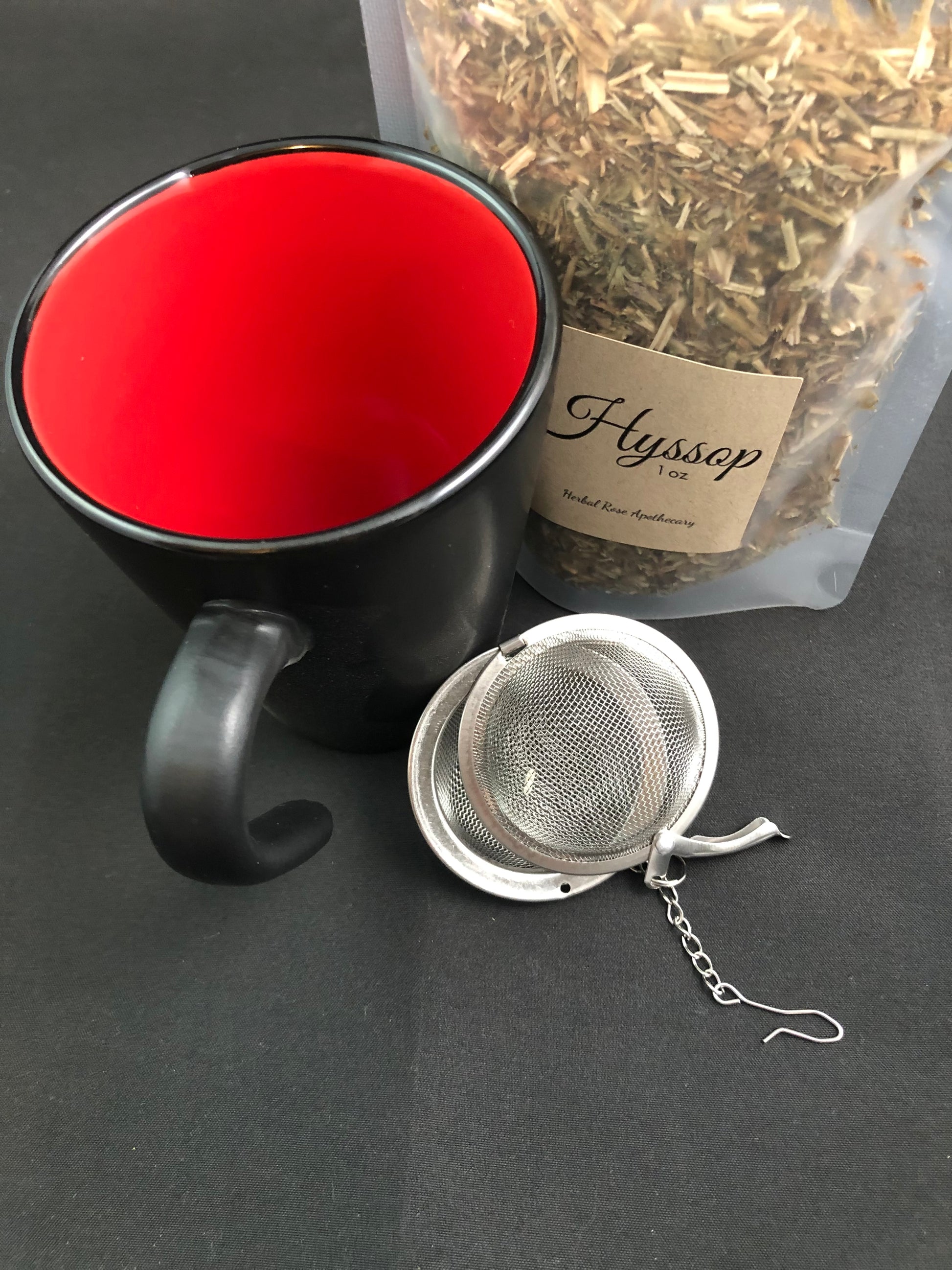 dried hyssop in a clear 1oz bag next to a black and red coffee mug and mesh tea infuser with a black background