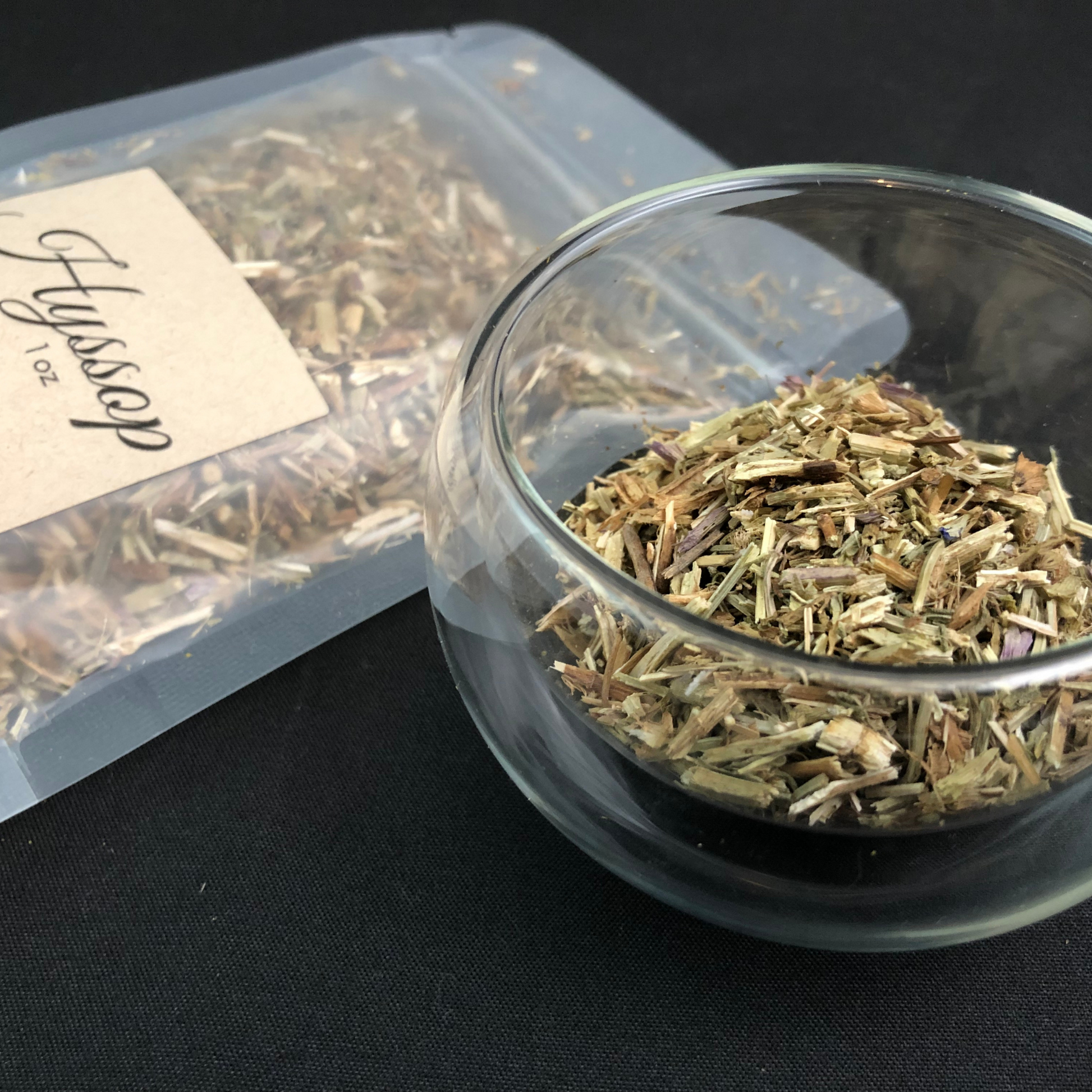 dried hyssop in a clear 1oz bag next to a clear glass cup of dried hyssop with black background