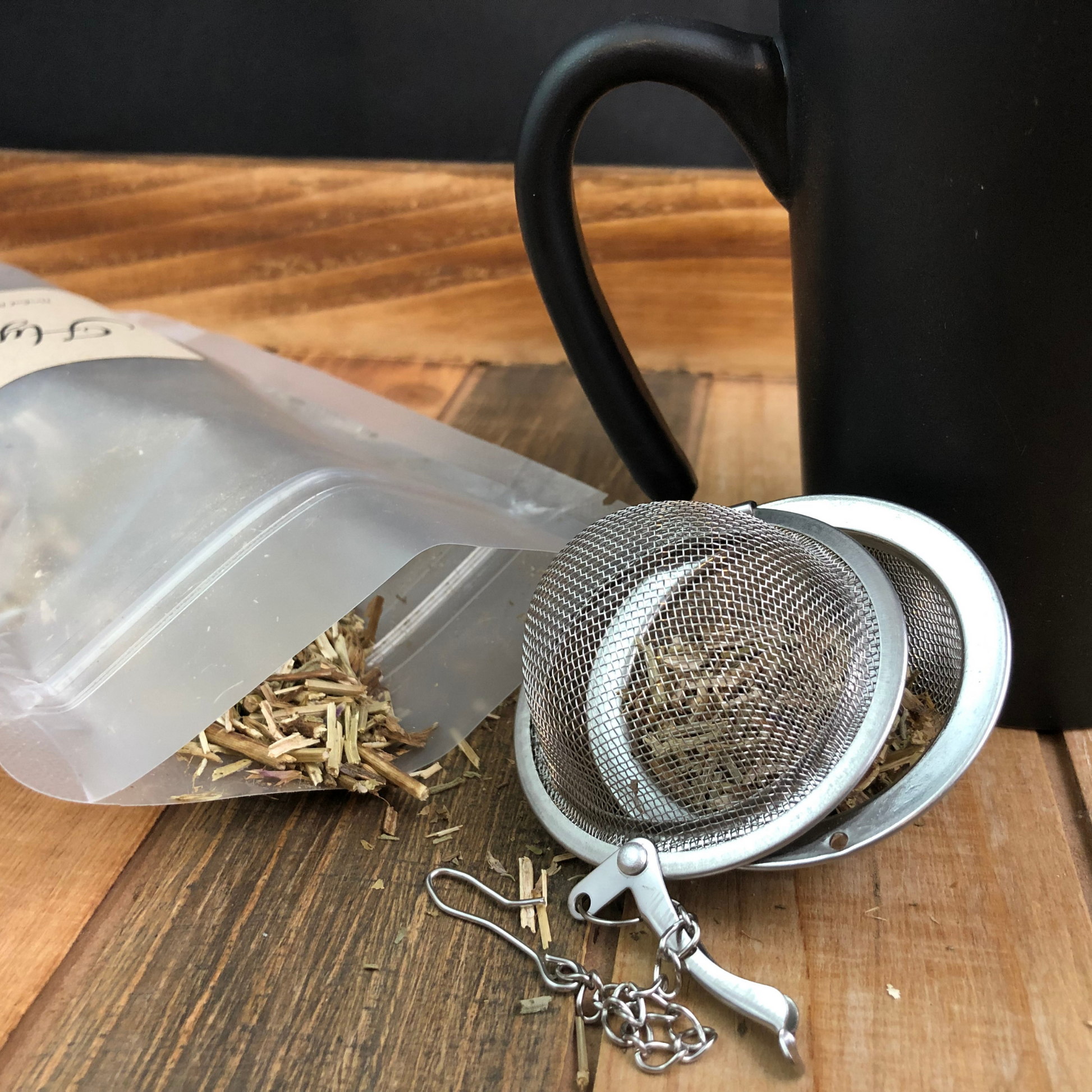 image of dried hyssop spilling out a clear bag next to a black and red coffee mug and mesh tea infuser on a wooden table with a black  background