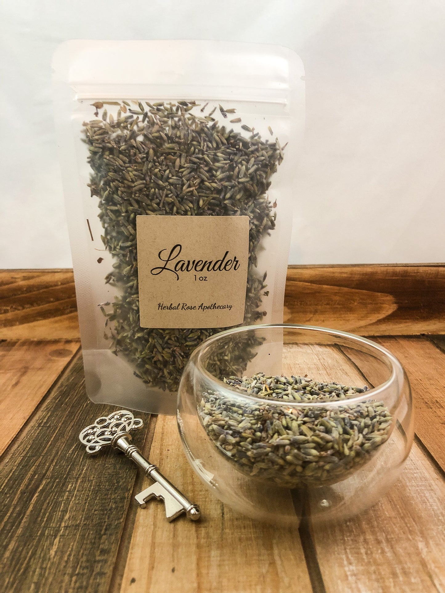 dried lavender in a clear 1oz bag next to a small clear glass cup of dried lavender, silver key laying in forefront, items on a wooden table with a white background