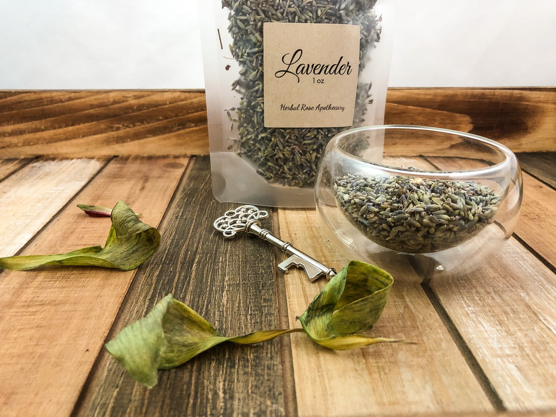 image of dried lavender in a clear bag, dried lavender in a small clear glass cup, a silver key and green leaves on wooden table with white background