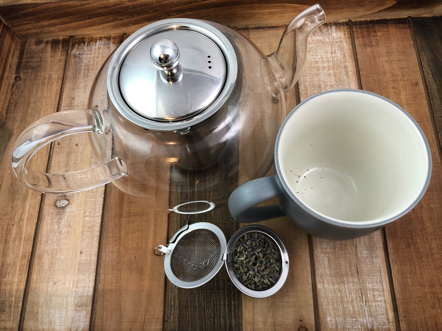 Ariel view of clear teapot, mesh tea infuser filled with dried lavender, next to a grey and white coffee mug on a wooden table as background