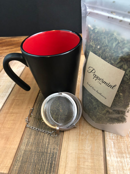 image of 1oz of dried peppermint in clear bag next to a black and red mug and mesh tea infuser with a black background, items on a wooden table