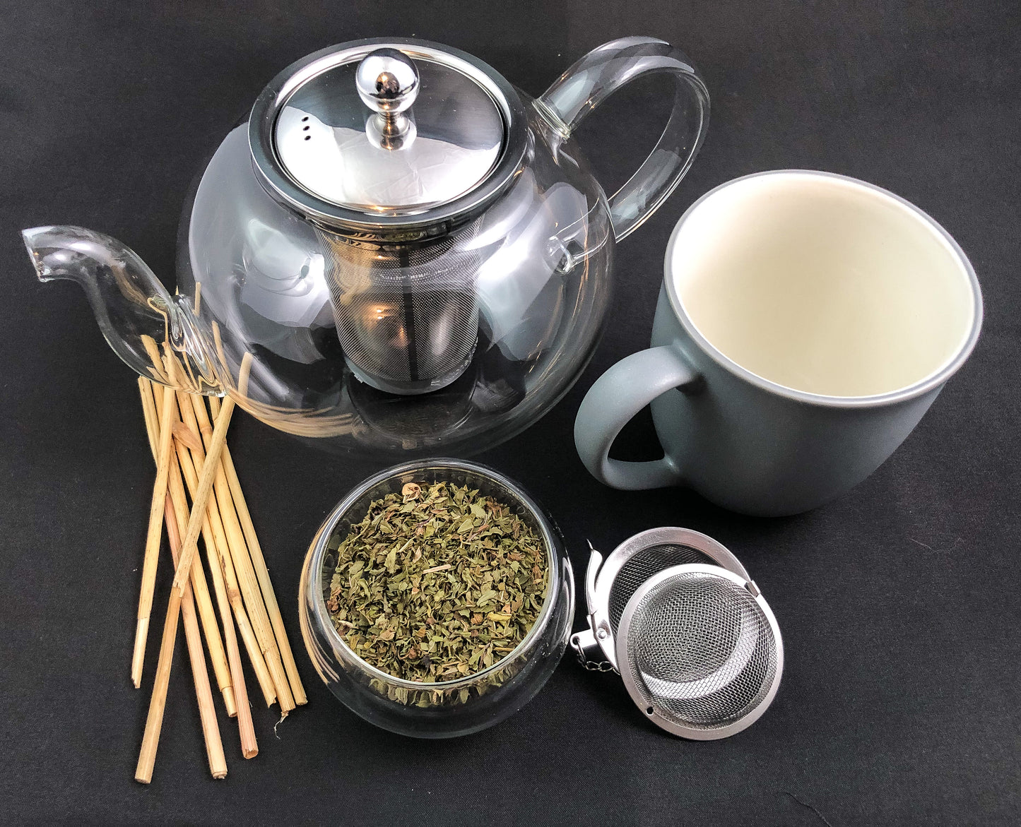 Ariel view of a clear teapot, grey coffee mug, mesh strainer, wooden sticks and a clear glass cup of dried peppermint with a black background