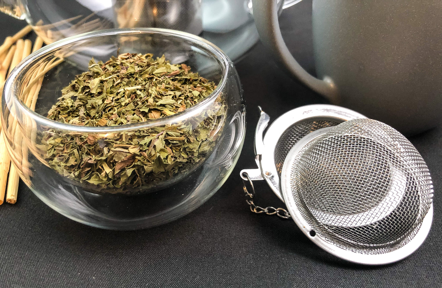 upclose image of dried peppermint in a clear glass cup next to a mesh tea infuser with black background and other items in the background