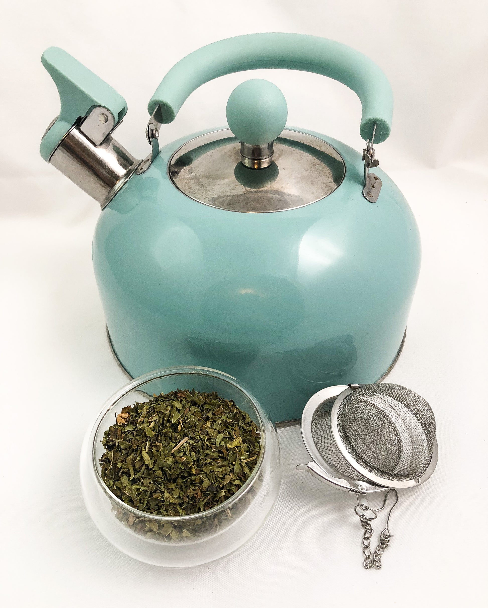 aqua colored teapot next to a small clear glass cup of dried peppermint leaves and a mesh tea infuser with white background