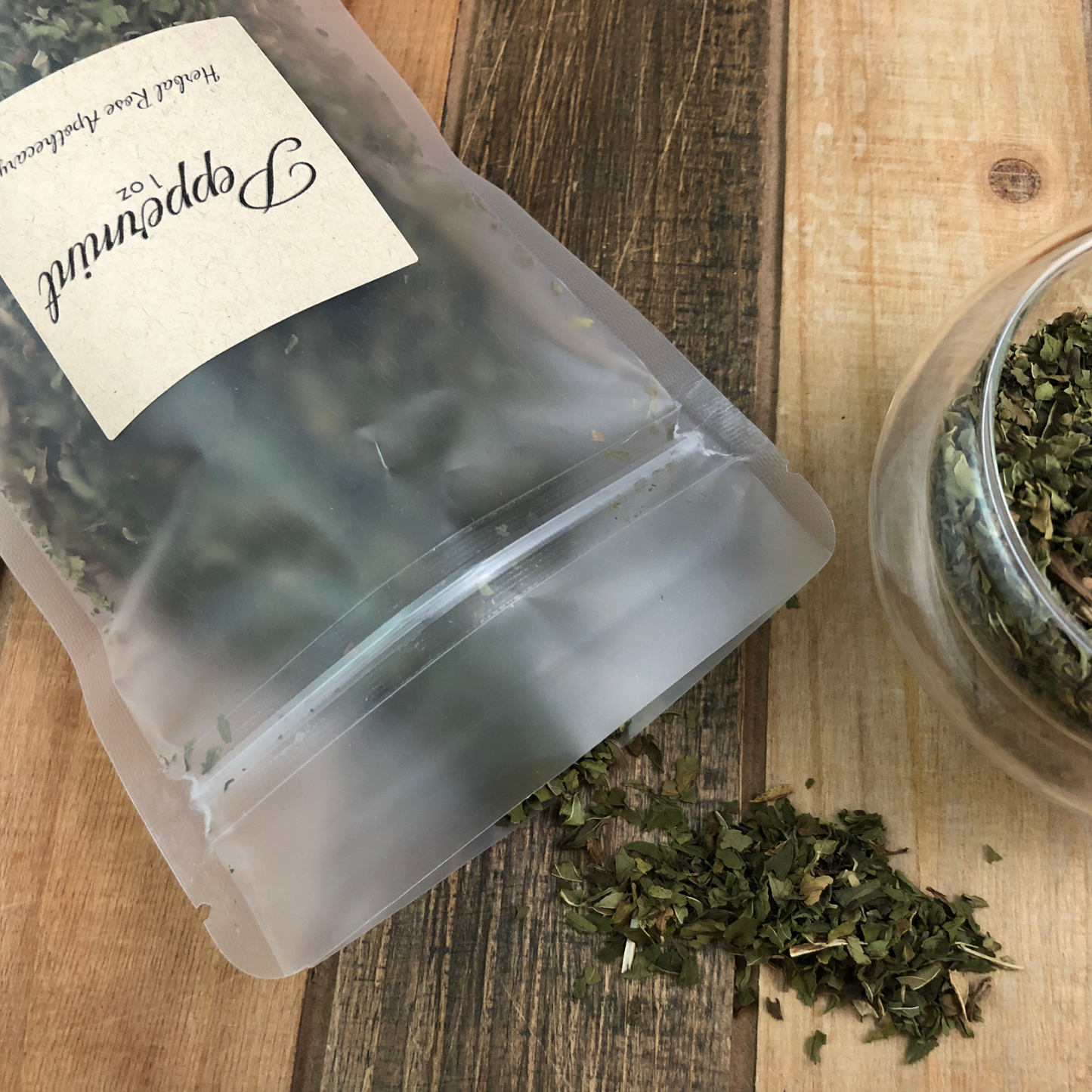 dried peppermint spilling out of a 1oz clear bag next to a clear glass cup of peppermint on a wooden table