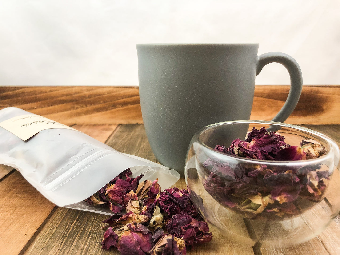 image of dried roses spilling out of a clear bag next to a small clear clear glass cup of dried roses and a grey coffee mug on a wooden table with white background