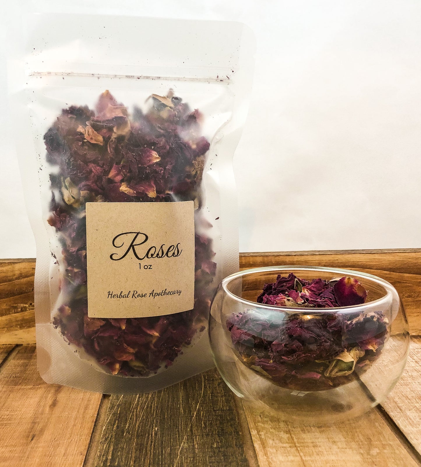 dried roses in a clear 1oz bag next to a small clear glass cup on a wooden table with a white background