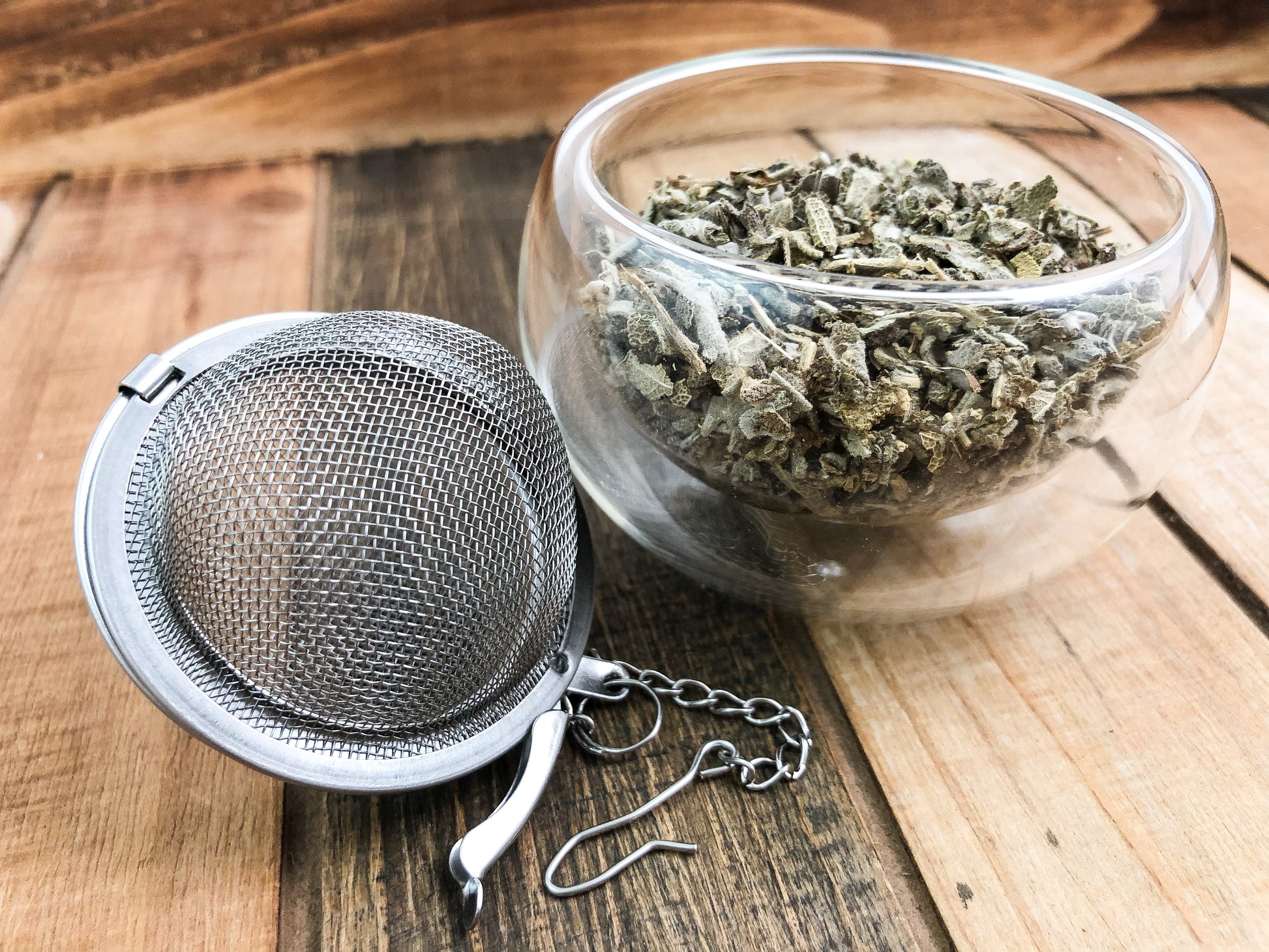 dried sage in a small clear glass cup next to a mesh tea infuser with a wooden table as the background