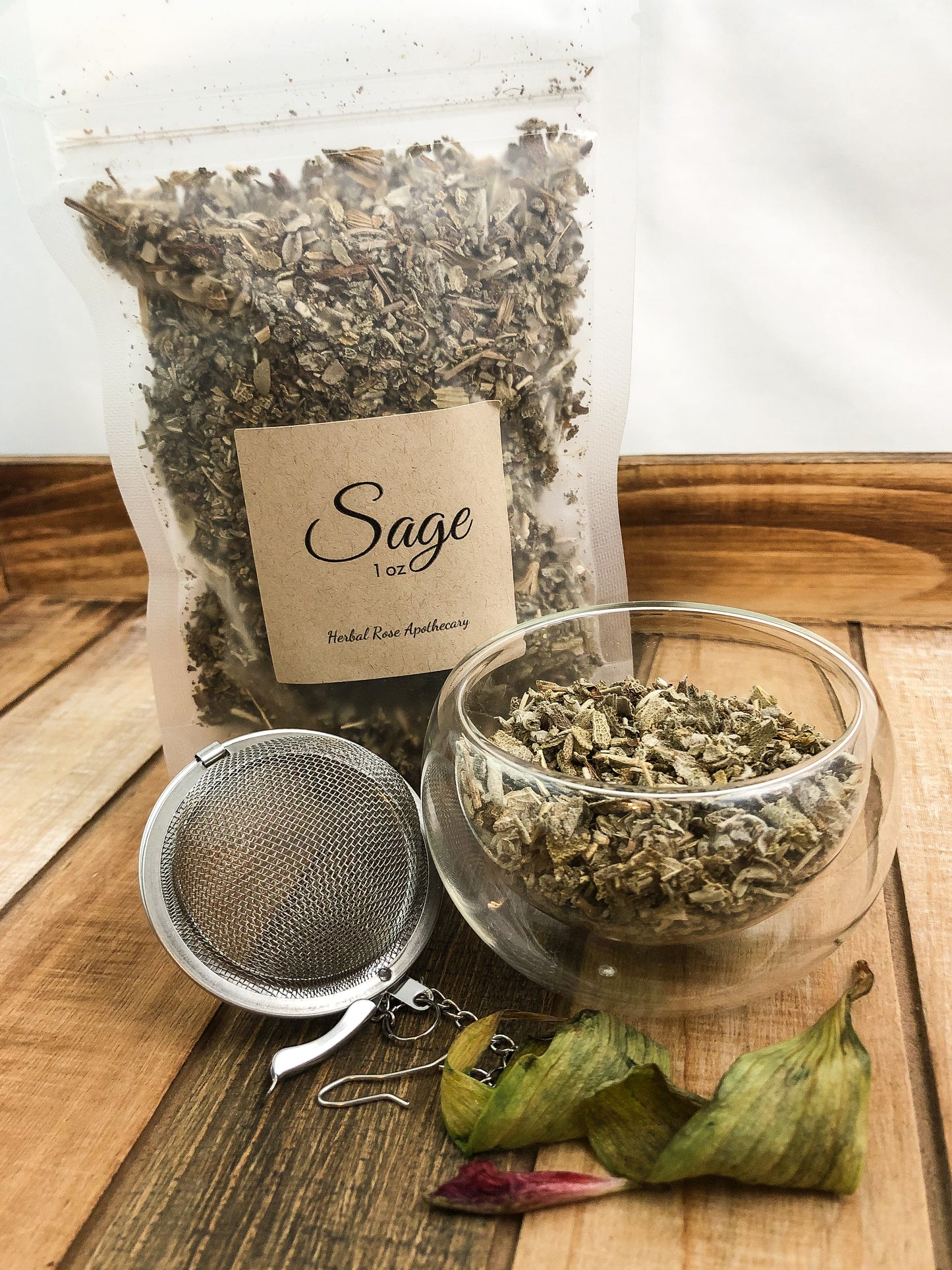 image of 1oz bag of dried sage next to dried sage in a clear glass cup and a mesh tea infuser with wilting flowers in front, white background and items on a wooden table