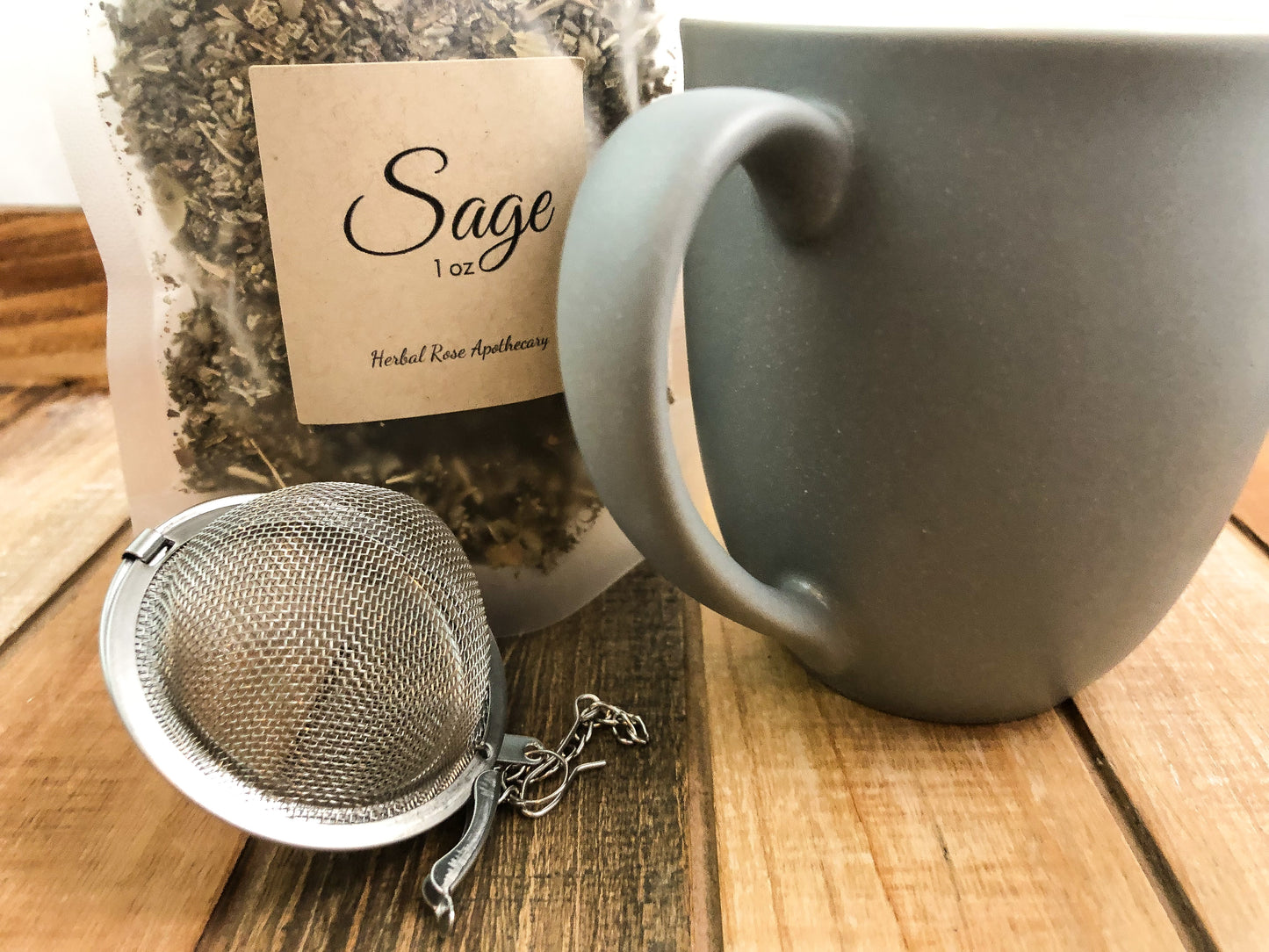 image of dried sage in a 1oz clear bag next to a grey coffee mug and mesh tea infuser on a wooden table with white background
