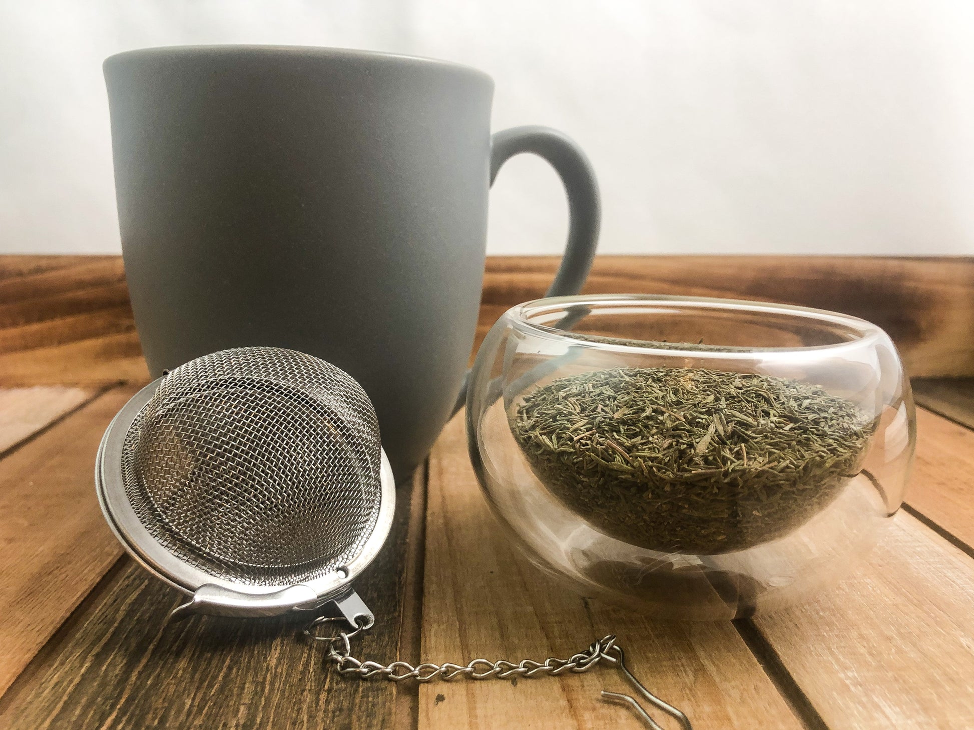 dried thyme in a clear glass cup next to a grey coffee mug and tea infuser on a wooden table with white background