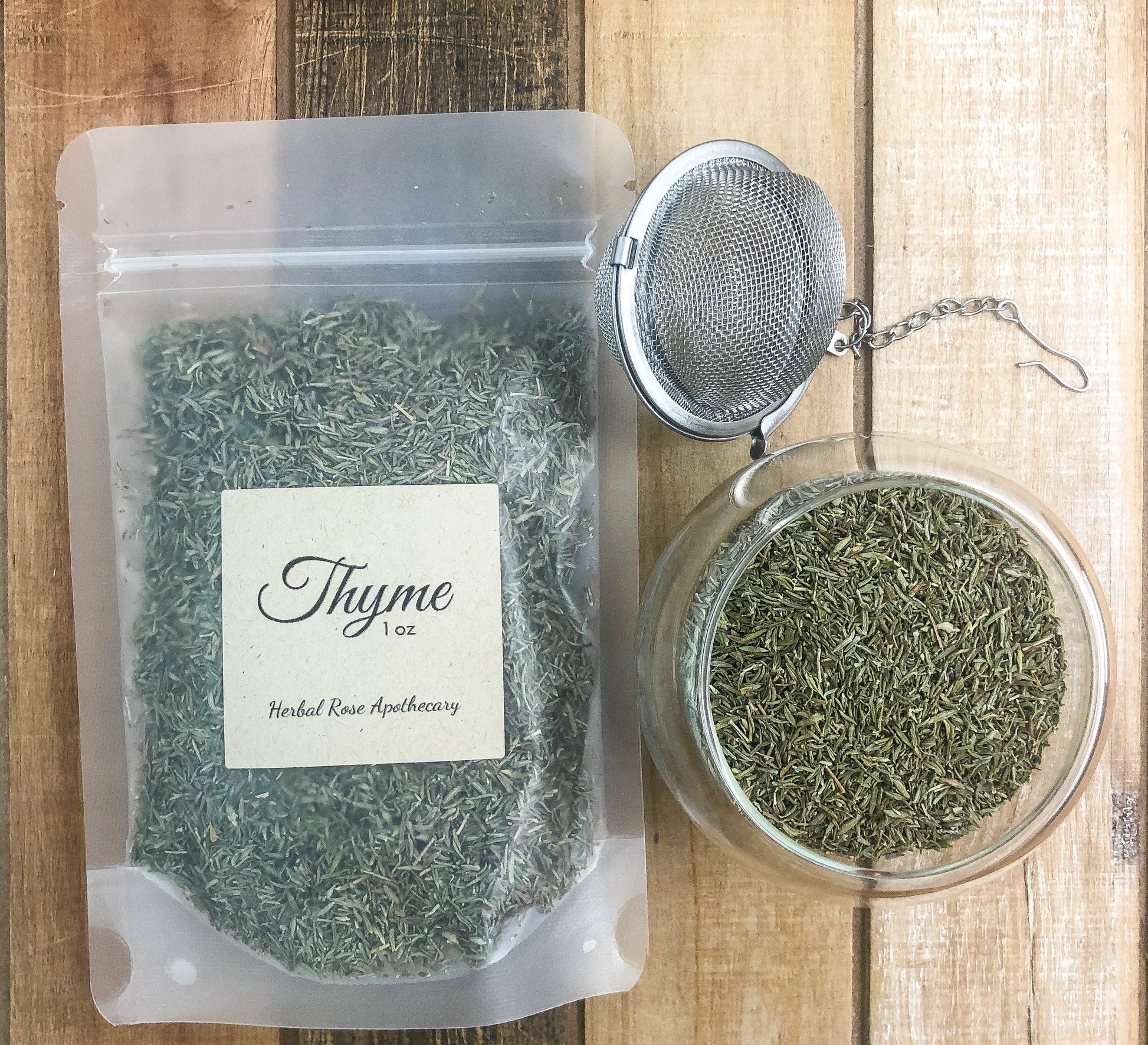 dried thyme in clear 1oz bag next to a mesh tea infuser and clear glass cup of dried thyme on a wooden table as background