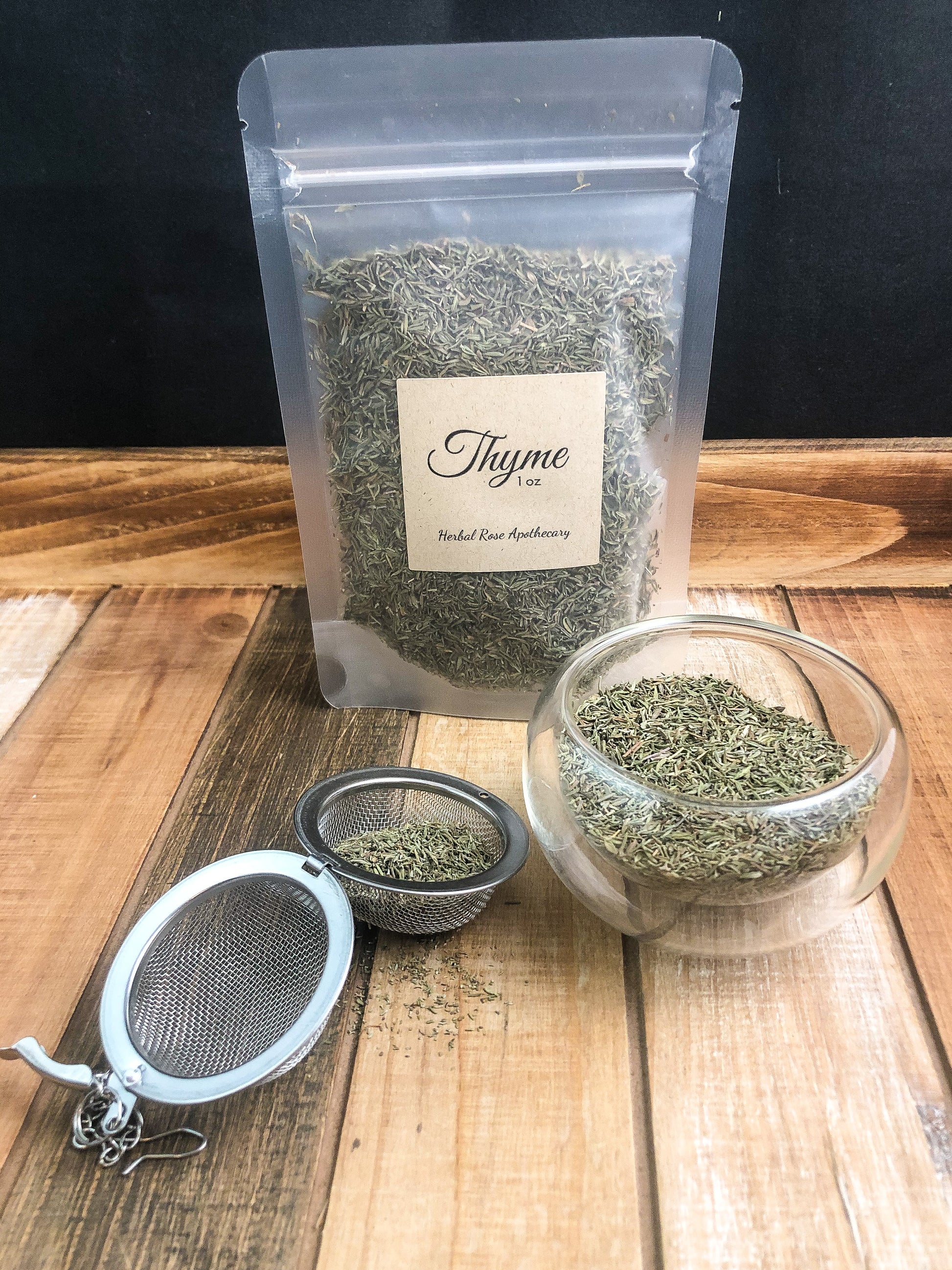 dried thyme in a clear 1oz bag next to dried thyme in a clear glass cup and thyme in mesh tea infuser on w wooden table as background