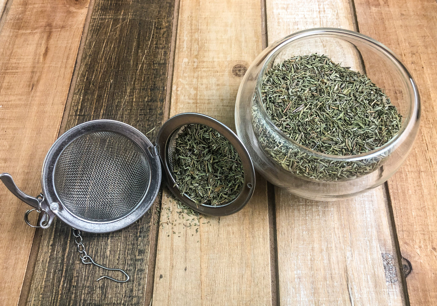 dried thyme in a clear glass cup next to a mesh tea infuser with dried thyme on a wooden table as background